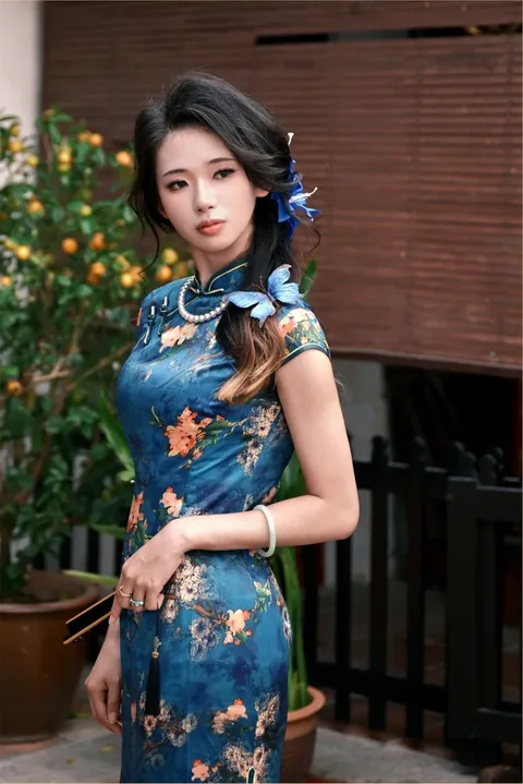 身穿蓝色印花旗袍、戴蝴蝶发簪的女子站在金桔树旁的庭院里