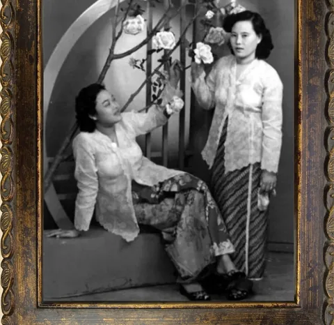 一张黑白老照片：两位女子身穿蕾丝可峇雅，站在照相馆的花枝布景前