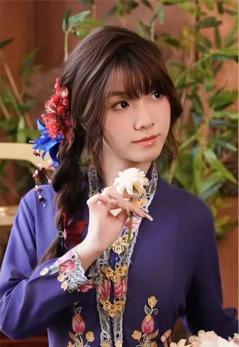 身穿紫色可峇雅的女子手执一朵菊花，身旁是室内绿植