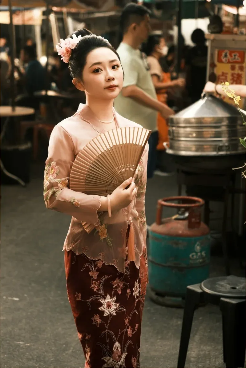 身穿粉色娘惹衫的女子手持折扇，站在傍晚的老巴刹摊档之间